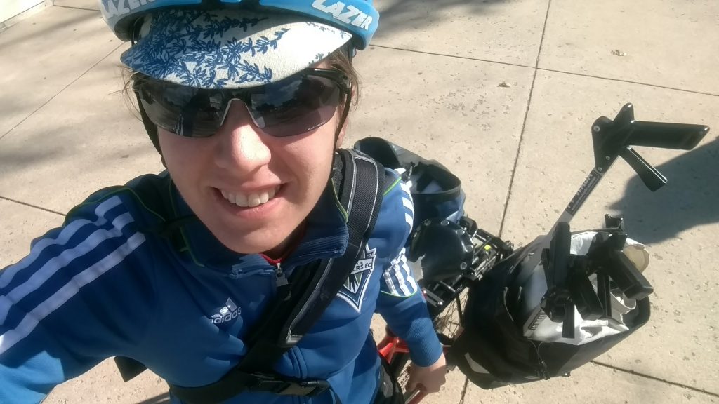 Selfie of a woman in a bike helmet, blue Seattle Sounders jacket and messenger bag. There are handles of four trash grabbers in the lower right sticking out of a bike pannier. 