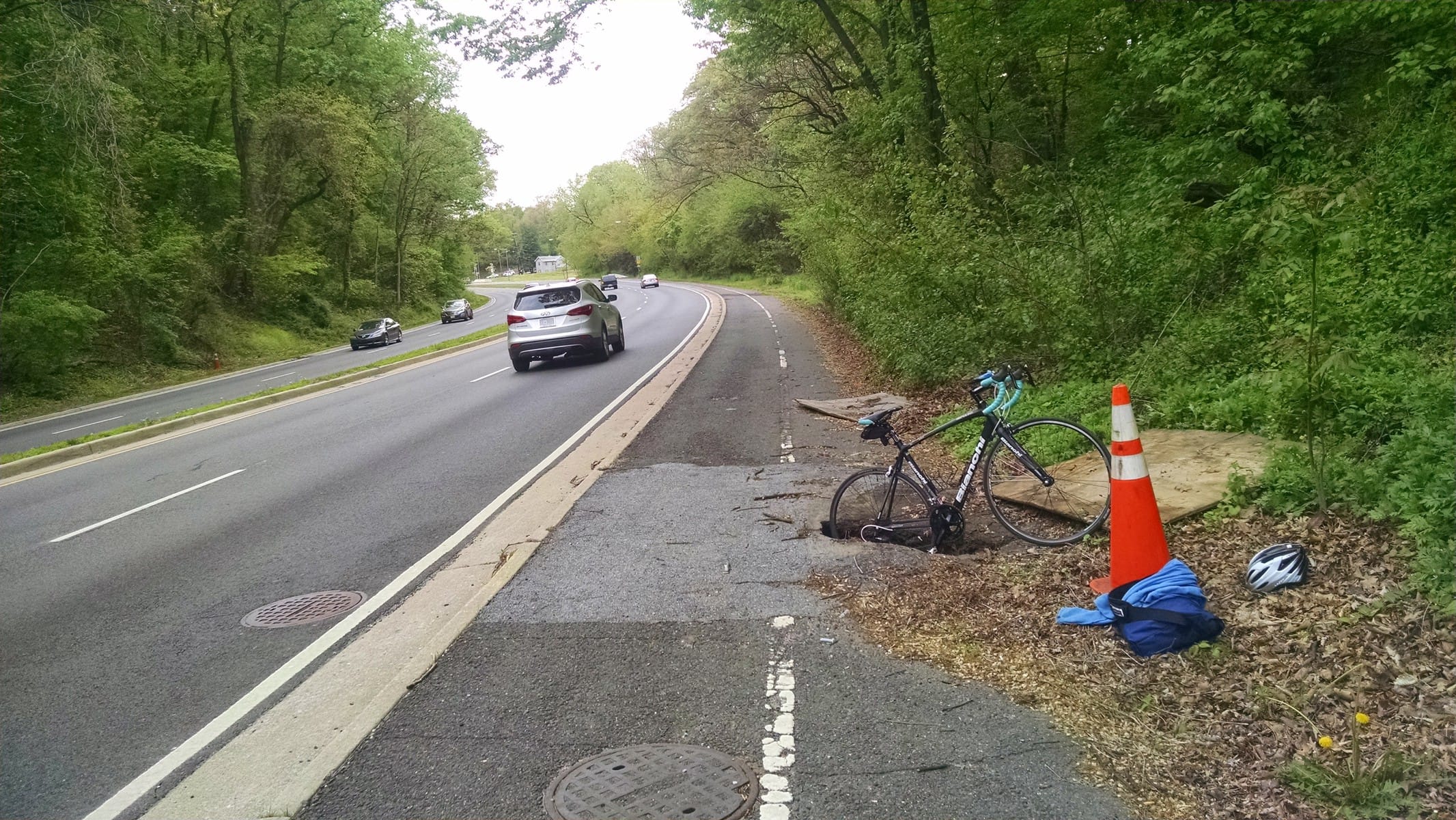 A trail in a state of disrepair next to a busy highway. A bicycle is shown with a wheel in a sinkhole, demonstrating the hole's size.
