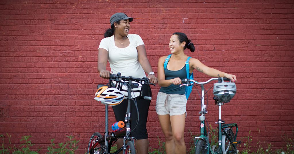 Two women with folding bikes laugh together in front of a red brick wall.