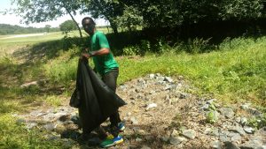 A Trail Ranger stands on some gravel holding a trash bag and trash grabber. He is smiling at the camera.