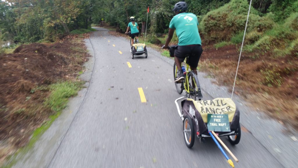 Two people bike away from the camera on a trail - they are both pulling trailers with brooms and are wearing Trail Ranger uniform shirts. 