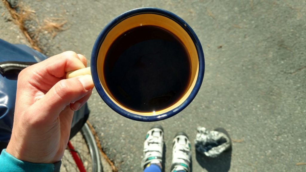 straight down shot of a metal yellow coffee mug filled with coffee. A white woman is holding it in her left hand and you can see her bike shoes on the ground in the distrance. 
