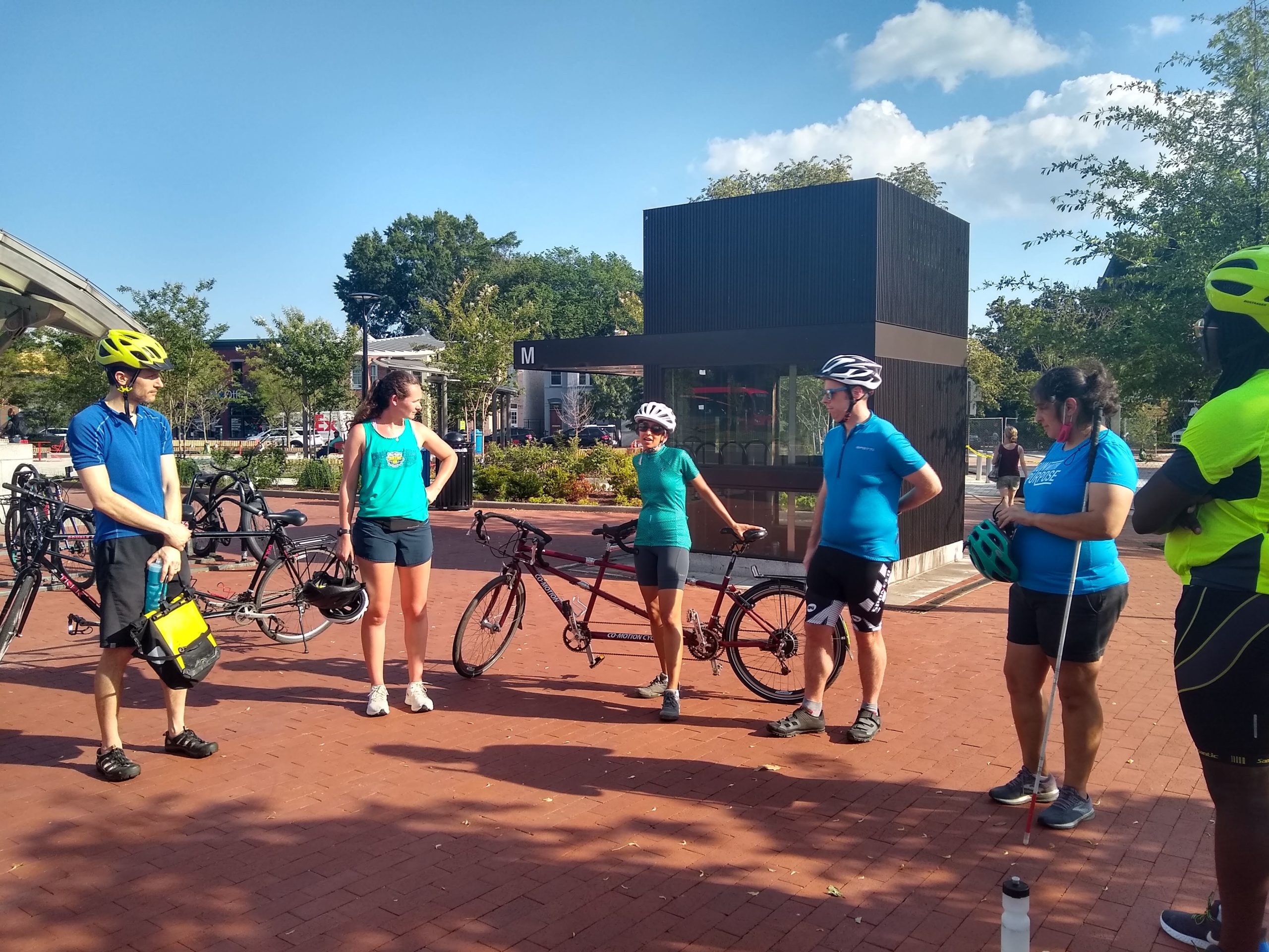 6 people are standing in a semicircle at Eastern Market Metro plaza in front of the elevator. Qudsiya is in the middle and speaking to the group. Everyone is spaced out, the sky is blue, it looks like a lovely day, and there are at least two tandems in the photo. A woman on the right has her cane in her hand and is holding a helmet. 