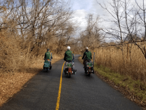 Three trail rangers in green jackets and on cargo bikes riding along on the Anacostia Riverwalk Trail in early Winter. The trail is damp and glistening and the vegetation on both sides is brown and the trees are leafless.