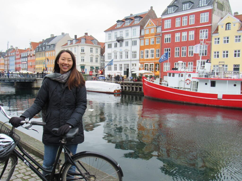 A photo of Michelle with a bike in front of a canal and colorful rowhouses in Copenhagen.